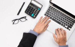A person in a suit works on a laptop at a white desk, searching for information about equity release mortgages in Canada, with a calculator, pen, and spectacles placed nearby.
